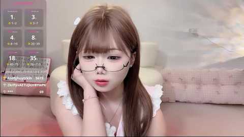 A video of a young East Asian woman with long brown hair, wearing glasses, lying on her stomach in a room with a pink couch.