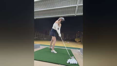 Video of a female golfer in a white sleeveless top, black skirt, and white cap, swinging a golf club on a green practice mat, under a white roof. Background features a dark sky, distant buildings, and golf course.