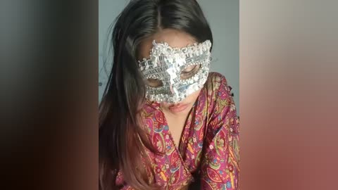 A video of a woman with long black hair, wearing a colorful, patterned blouse and a decorative silver masquerade mask with intricate designs, leaning forward against a blurred background.