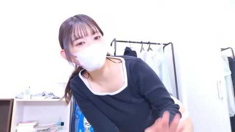 Video of an Asian woman with long dark hair, wearing a white face mask, black long-sleeved shirt, and white bra, in a minimalist room with clothes hanging on a rack.