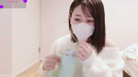 Video of an East Asian woman with straight, shoulder-length black hair, wearing a white mask and fluffy white sweater. She's holding a light blue drink, in a simple, brightly lit room with a bed in the background.
