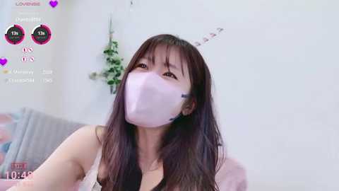 A video of an Asian woman with long, dark hair and a pink surgical mask, wearing a black top, taken in a room with white walls and a hanging plant.