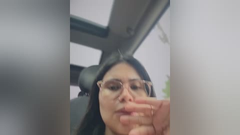 A blurry, close-up video of a woman with long black hair and glasses, seen from the car window, with a hand near her mouth.