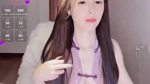 Video of a young, pale-skinned, slender Asian woman with long, straight, dark brown hair, wearing a sheer, lavender dress with purple ties, sitting on a white fur rug. Background features a white wall and a purple heart icon.
