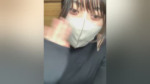 Video of a person with a light skin tone wearing a gray face mask, dark long hair, and a black top, standing in a room with a beige wall and wooden blinds in the background.