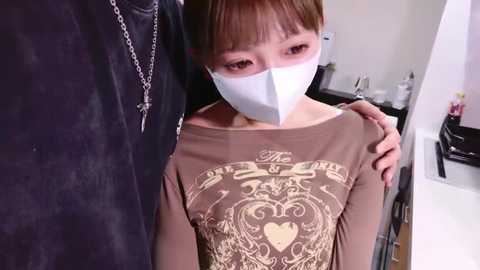 Video of an Asian woman with short brown hair, wearing a face mask, pulling down her brown T-shirt revealing an ornate heart design. Background shows a bathroom with toiletries.