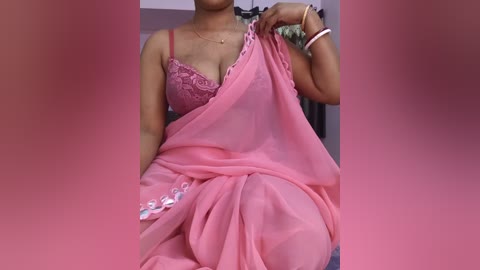 Video of a woman in a pink, see-through saree with floral embroidery, holding the fabric. She wears a matching pink bra. Background shows a partially visible room with a grey carpet and a black and white garment.