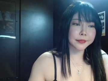 Media: Video of a young Asian woman with long black hair, fair skin, and light makeup, wearing a black spaghetti-strap top, standing indoors against a dark, dimly lit background.