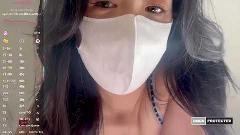 Video of a woman with long black hair, wearing a white face mask, black top, and visible facial skin. Background includes a video call screen with a \"Protected\" label.