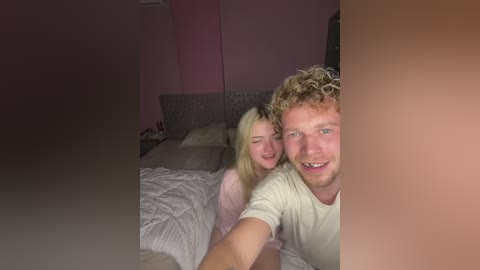 A video of a young couple in a dimly lit bedroom, with pink walls and a bed. The man has curly blonde hair, and the woman has straight blonde hair. Both are smiling.