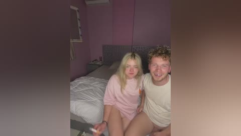 Video of a young couple, a blonde woman in a pink pajama top and a curly-haired man in a beige t-shirt, sitting on a bed in a pink bedroom.