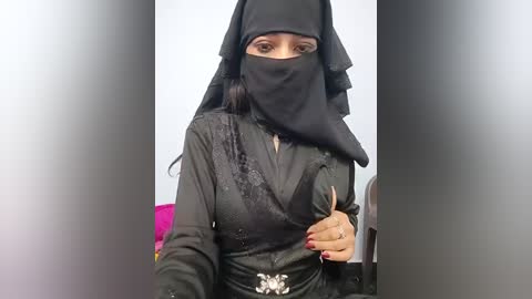 Video of a woman in a black burqa, with a veil covering her face, wearing a black dress with intricate embroidery and silver belt buckle. She stands indoors, with a blurred background.