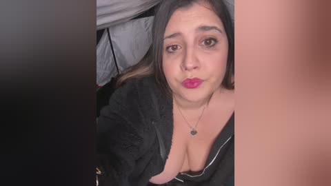 Video of a light-skinned woman with long dark hair, wearing black clothing, lying on a bed, with a prominent cleavage and a necklace, partially visible.