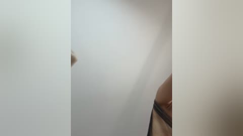 A video of a naked woman's face and right breast, with a small hole in the white wall to her left. Her skin tone is light brown, and her hair is partially visible. The background is plain white.