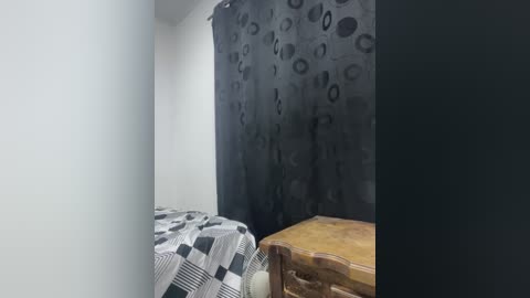 Video of a minimalist bedroom with a bed covered in a black-and-white checkered blanket, a wooden bedside table, and a dark gray wall with abstract black patterns.
