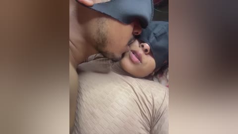 Video of a man in a black face mask passionately kissing a woman with pink lipstick, both wearing light-colored tops, in a dimly lit room.