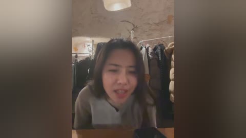 Video of a young Asian woman with shoulder-length dark hair, wearing a beige top, laughing while looking at a phone, surrounded by hanging clothes in a dimly lit, cozy bedroom.