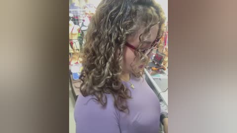 Video of a young woman with curly, light brown hair, wearing red-framed glasses and a light purple top, standing in a brightly lit, cluttered room with colorful posters on the walls.