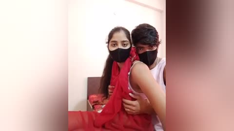 Video of a woman in a red saree and face mask, being held by a man in a white tank top, in a dimly lit room with beige walls.