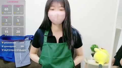 Video of an East Asian woman with long black hair wearing a green apron over a black shirt, standing in a kitchen with a blue and white striped towel and a yellow toy on a white shelf.