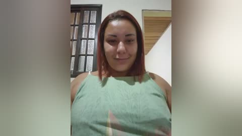 Video of a smiling woman with shoulder-length red hair, wearing a green sleeveless top, standing in a modern, brightly lit room with a grid-patterned window and wooden door in the background.