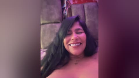 A video of a smiling, topless woman with long black hair, lying on a gray bed, surrounded by a dimly lit, purple-hued room.