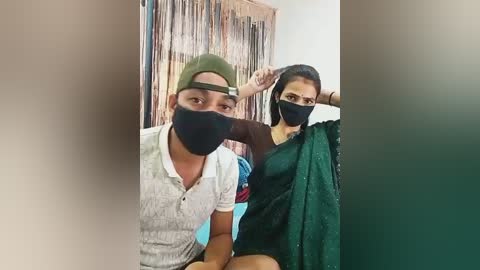 Video of two South Asian individuals wearing black face masks, one in a green cap and white shirt, the other in a green sari, sitting on a bench in a room with a decorative wall hanging.