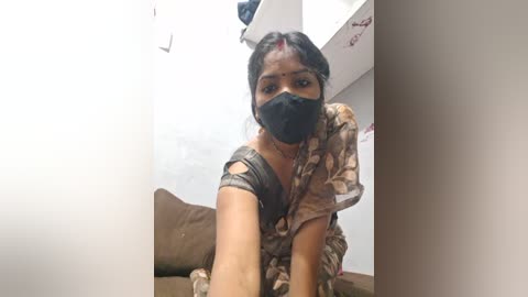 Video of a South Asian woman with medium-dark skin, wearing a black mask, floral saree, and red bindi. She's kneeling on a brown sofa in a dimly lit room with white walls.