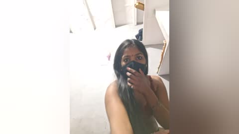 A video of a nude South Asian woman with long black hair, partially covering her face, taking a selfie in a bright, cluttered room with white walls and furniture.