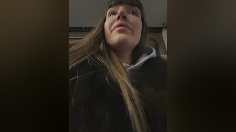 A video of a young woman with long, straight brown hair and light skin, wearing a black coat and a gray hoodie, standing indoors with a neutral background.