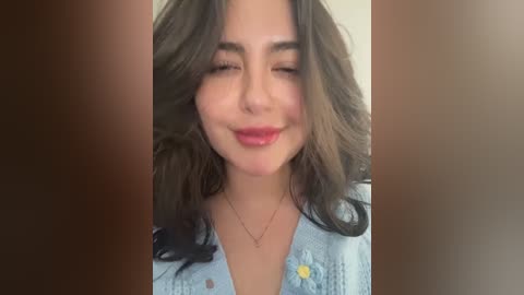 A video of a young woman with long, wavy brown hair, fair skin, and full lips, wearing a light blue denim jacket with a yellow button. She has a slight, content smile.