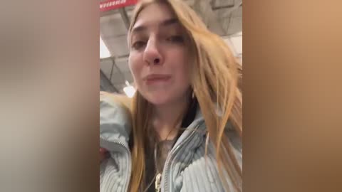 A video of a young woman with long, blonde hair, wearing a light blue denim jacket, taken in an indoor setting with blurred background.