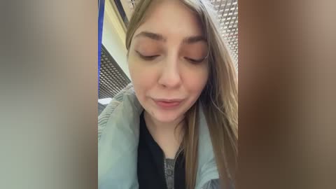 Video of a young woman with light skin and straight, light brown hair, wearing a light blue jacket over a black top, eyes closed, smiling, taken indoors with a tiled wall and metal ceiling in the background.