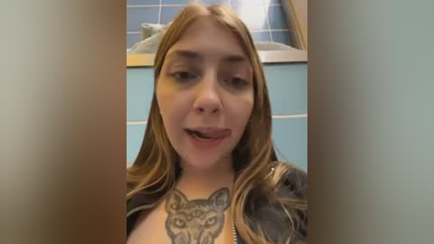 Video of a young woman with long, straight brown hair and fair skin, wearing a dark jacket, standing in a tiled bathroom with blue and white tiles. She has a large, detailed wolf tattoo on her chest.