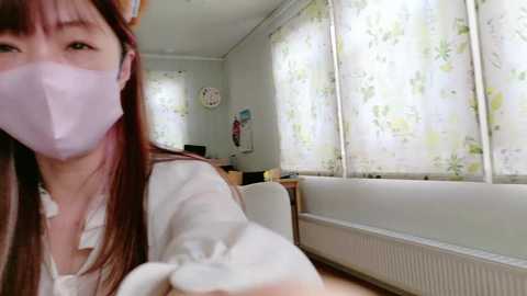 Video of a young Asian woman with long brown hair, wearing a white mask and floral blouse, sitting in a sunlit, white-walled room with floral-patterned curtains and a white radiator.