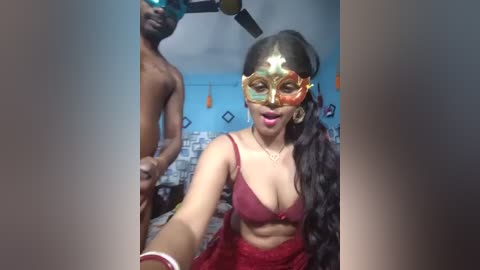 Video of a South Asian woman in a red lace bra, gold mask, and long black hair, posing indoors with a shirtless man in the background.