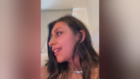 A video of a young woman with long, wet hair, light skin, and dark eyes, smiling slightly, wearing a white top, in a bathroom with blurred, neutral-toned walls.