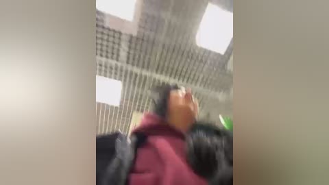 A blurry video of a person with short black hair in a red hooded jacket, lying on a black car seat in an indoor, grid-patterned, dimly lit room.