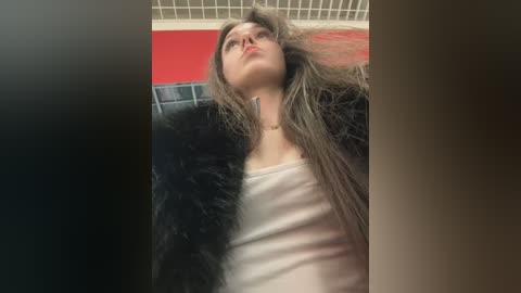 Video of a young woman with long, tousled hair, wearing a white tank top and black fur coat, leaning against a red wall in a dimly lit room.