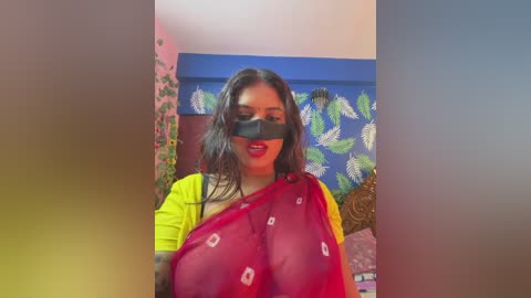 Video of a dark-skinned woman with long black hair, wearing a bright yellow top, a red saree, and a black blindfold, standing in a room with blue curtains and green leaf patterns.