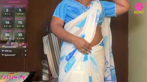 Video of a South Asian woman in a blue and white sari, holding a striped bag, standing in a kitchen with a calendar displaying social media followers.