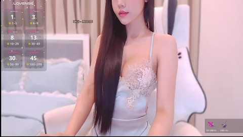 Video of an Asian woman with long, straight black hair, fair skin, and wearing a revealing, light blue, lace-trimmed lingerie top, sitting on a bed with white sheets and a white headboard.