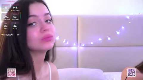 A young woman with long dark hair and fair skin, wearing a white spaghetti-strap top, sits on a bed with a beige padded headboard. A live stream with a chat window and heart emojis overlays the image.