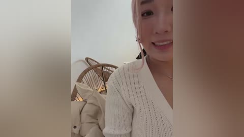 A video of a smiling Asian woman with fair skin, wearing a white knit sweater, standing indoors beside a wicker chair with a beige cushion.