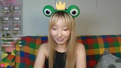 Video of a smiling Asian woman in a black tank top, wearing a green frog headband with large eyes and a crown, sitting on a colorful plaid couch.