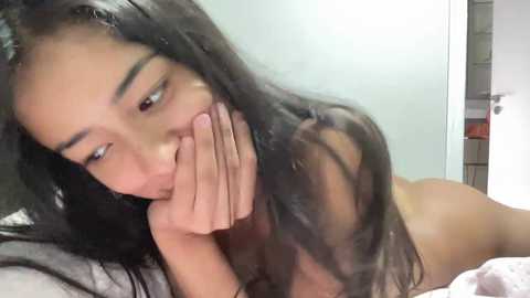 A video of a young, naked woman with long black hair, lying on her stomach on a bed, covering her mouth with her hand. Her skin is light brown. The background is blurred, showing a white door and a wall.