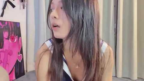 Media: Video of a young woman with long, dark hair, wearing a sleeveless top, speaking in a room with a pink poster and beige curtains.