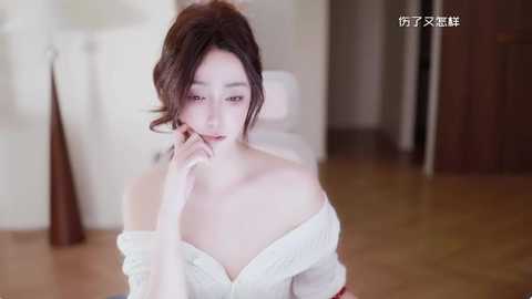 A video of a fair-skinned Asian woman with voluminous dark hair, wearing a white off-shoulder top, in a softly lit, modern room with wooden floors and neutral decor.