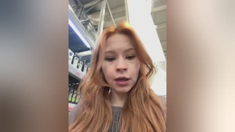 A video of a young woman with long, wavy, reddish-brown hair, wearing a grey knit sweater, standing indoors. The background features a blurred, modern, high-ceilinged space with white beams and fluorescent lighting.