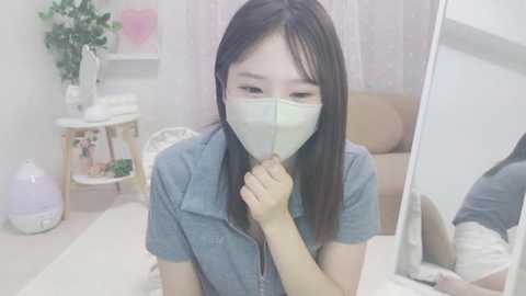 Video of an East Asian woman with long black hair and fair skin wearing a face mask, blue shirt, and seated at a white table in a minimalist room with beige walls and a mirror.